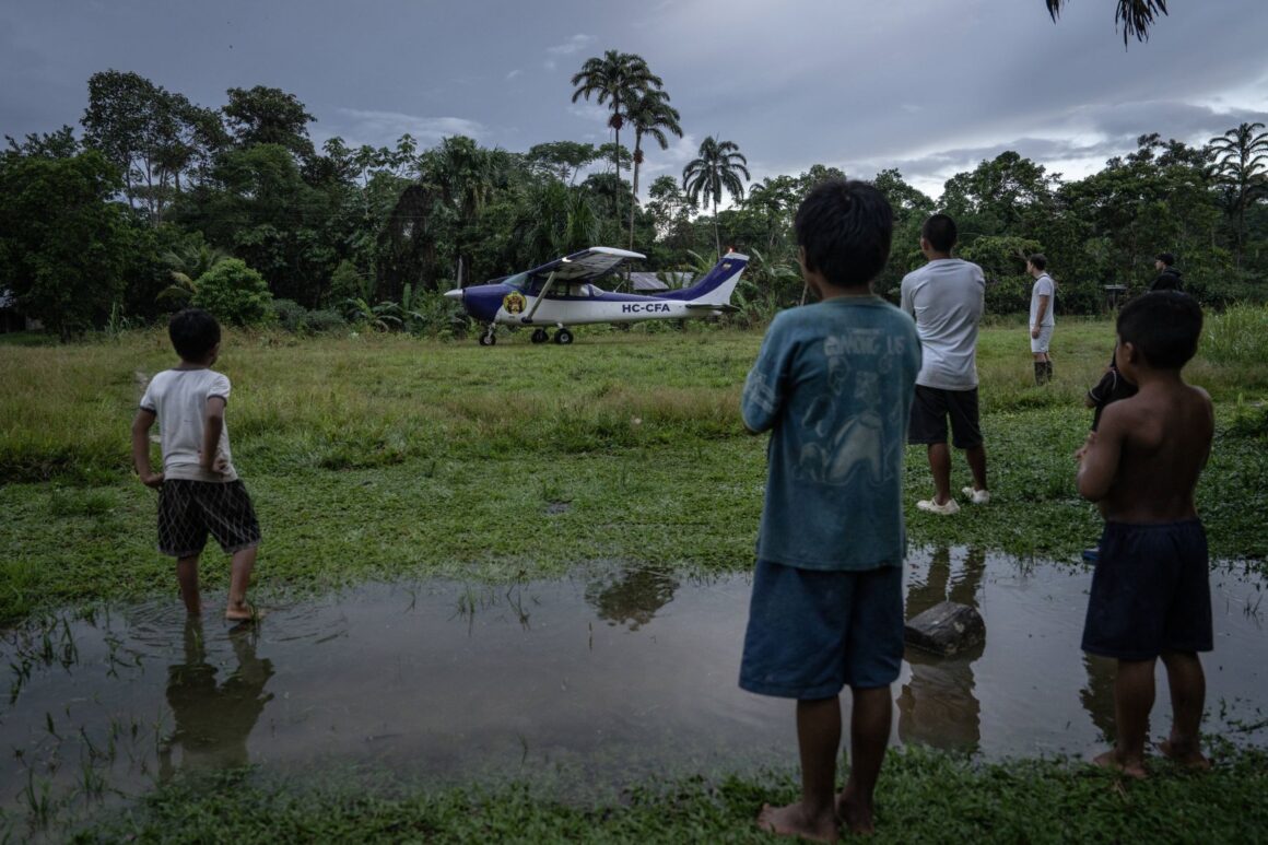 Waorani children stand near a dirt airstrip in the Amazon, watching a small plane land.