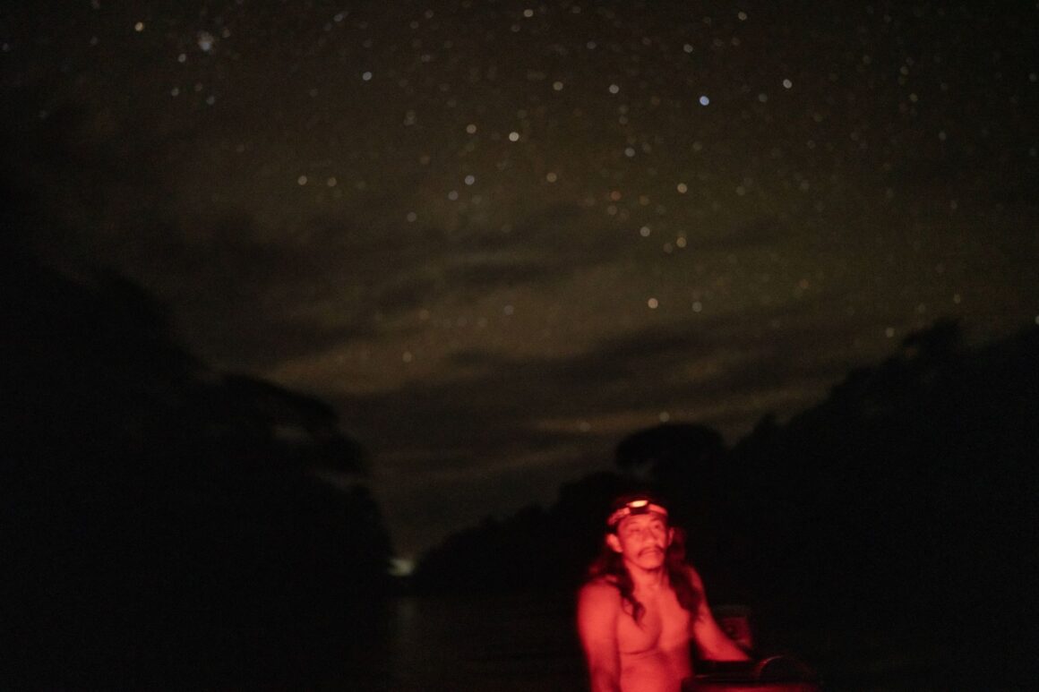A Waorani man paddling a wooden canoe on a river at night in the Amazon rainforest.