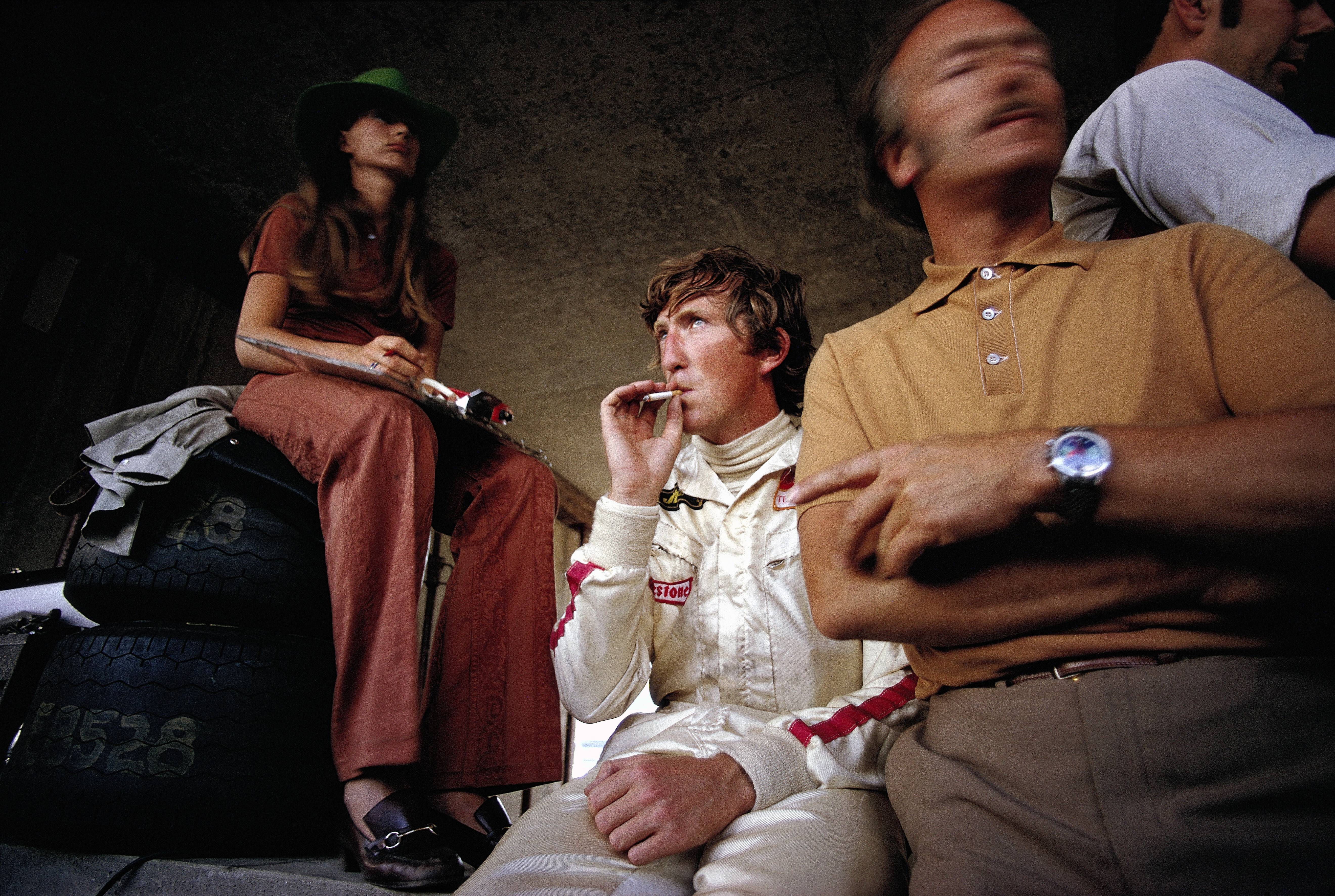 IMAGO / Motorsport Images | Jochen Rindt smokes a cigarette in the pits alongside his wife Nina and Lotus team boss Colin Chapman during the Austrian GP at Red Bull Ring. 16 August, 1970.