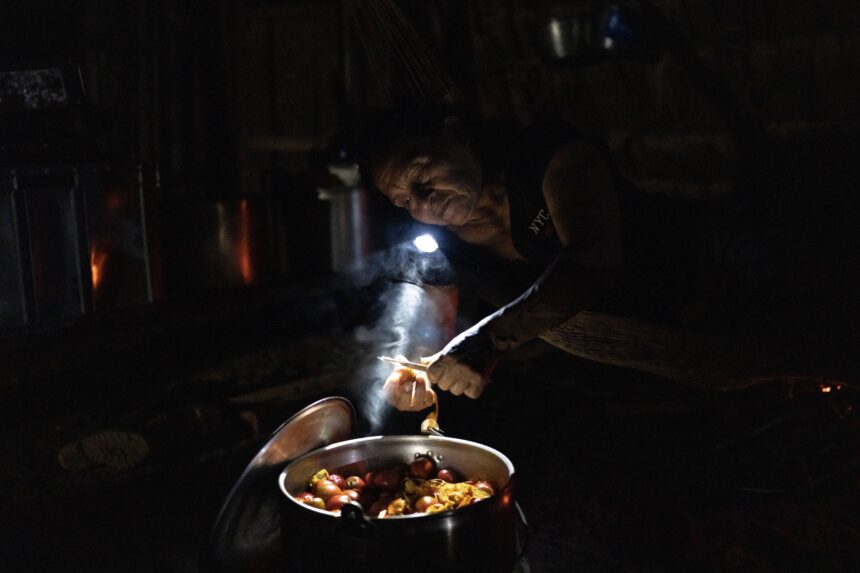A Waorani man cooks in dim light, assisted by someone holding a light source, while preparing a meal in the Amazon.